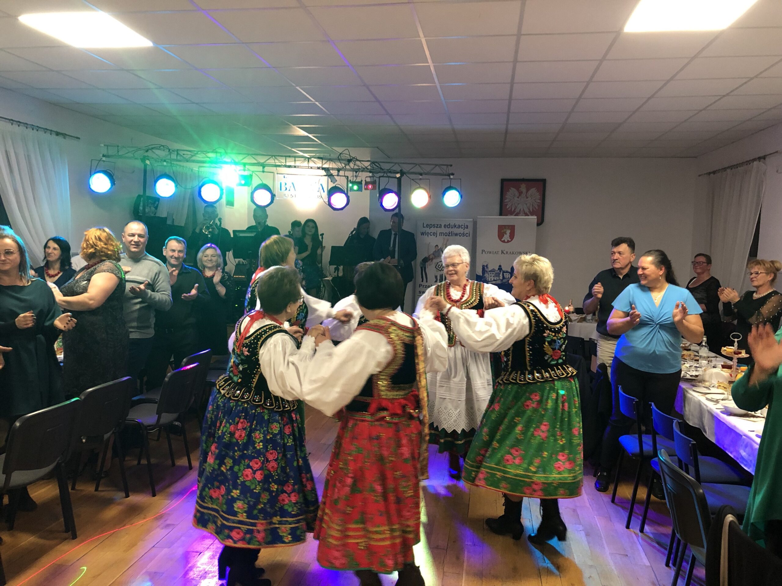 zdjęcie tańczących Pań w strojach krakowskich w tle sala, ludzie stoją i klaszczą