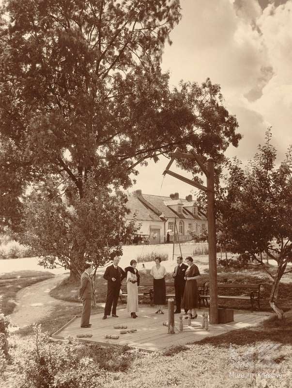 Fotografia czarno-biała. Sanatorium w Batowicach. Kręgielnia w parku. Na placyku pomiędzy drzewami stoi sześć osób elegancko ubranych, w tym trzy kobiety. Przed nimi rozstawione drewniane kręgle. W głębi zabudowania.